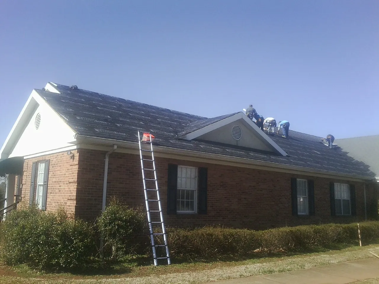 Crew tearing shingles off a brick ranch roof with a ladder set — Metal Roof Repair work by our crew in Troy